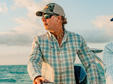 Man wearing a plaid shirt, sunglasses, and camo hat steering a boat on the ocean during sunset.