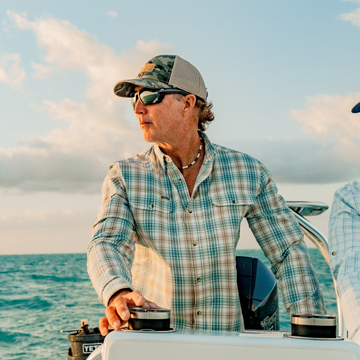Man wearing a plaid shirt, sunglasses, and camo hat steering a boat on the ocean during sunset.