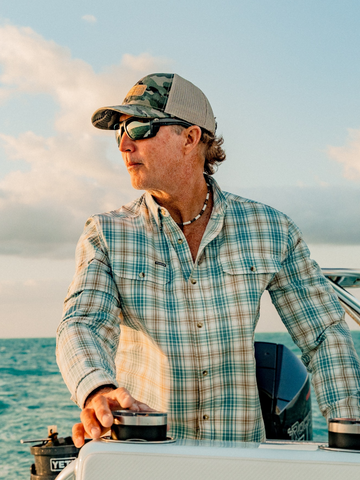 Man wearing a plaid button-up shirt and camouflage cap steering a boat on the ocean at sunset