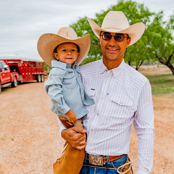 Man and child wearing cowboy hats and western shirts standing outdoors on a gravel path near trees and vehicles.