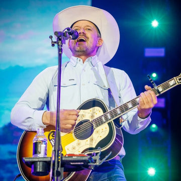 Man wearing a white cowboy hat and light blue shirt playing an acoustic guitar and singing into a microphone on stage
