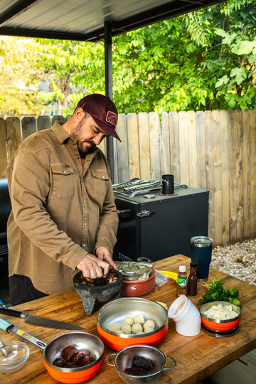 Man in a brown shirt prepares food outdoors beside a wooden table with bowls and cookware