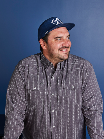 Man wearing a patterned button-up shirt and a navy blue baseball cap smiling against a blue background