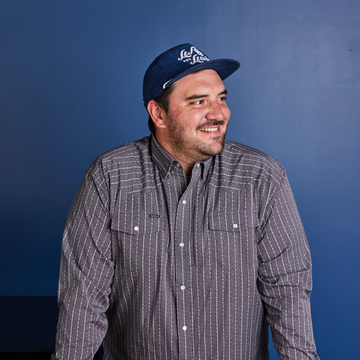 Man wearing a patterned button-up shirt and a navy blue baseball cap smiling against a blue background