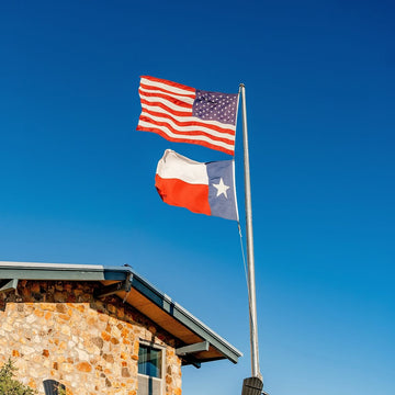 American and Texas flags flying beside a rustic stone building under a clear blue sky