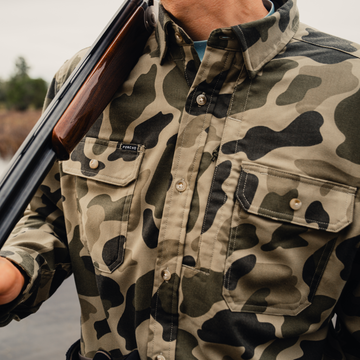 Man holding shotgun wearing camo button down shirt