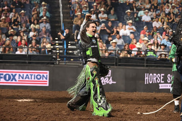 Performer in a black and neon green fringed outfit raises a fist in a dirt arena before a cheering crowd