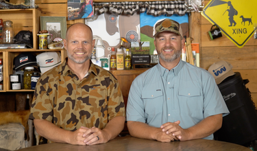 Two smiling men in button up shirts pose behind a wooden counter in a rustic outdoor themed room