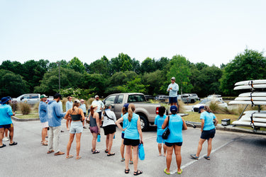Group of people gather near stacked kayaks and pickup trucks at a wooded outdoor launch area