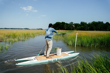 Man in a blue outdoor shirt standing on a paddleboard in a grassy marsh wetland while fishing