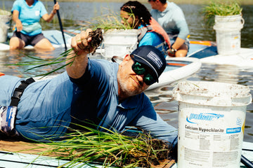 Man in blue outdoor shirt and sunglasses holds aquatic plants beside a boat with buckets and people nearby