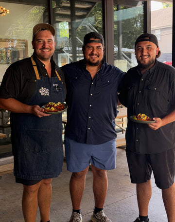 Three smiling men in dark shirts and aprons hold plates of food indoors near large windows