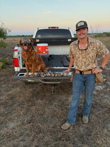 Man wearing a camouflage outdoor shirt beside a dog and pickup truck with fish in the truck bed