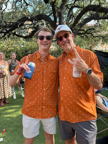 Two men wearing matching orange patterned outdoor shirts under a large oak tree in a sunny park