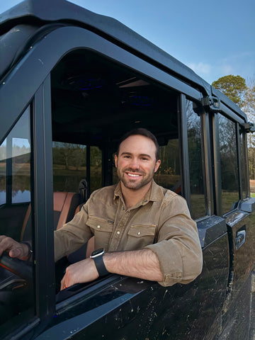 Man smiling inside off-road vehicle wearing brown button-up shirt outdoors
