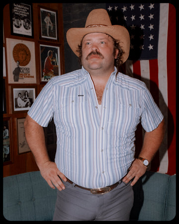 Man wearing a light blue striped short sleeve shirt and cowboy hat in front of an American flag