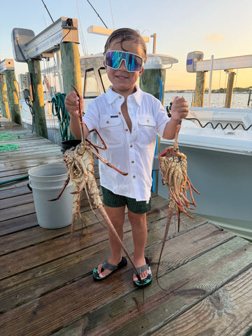 Child wearing sunglasses and a white button-up shirt holds freshly caught lobsters on a fishing dock