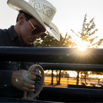 Man in a cowboy hat and dark shirt grips a rope beside a fence at sunset