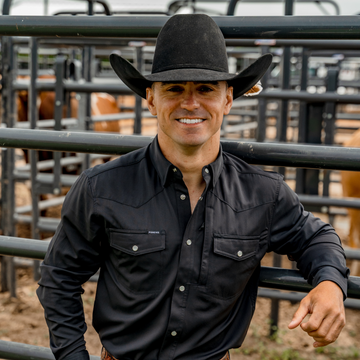 Man wearing a black western shirt and black cowboy hat leaning on metal fence in a livestock pen outdoors