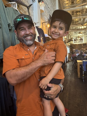Smiling man in an orange shirt holding a young boy inside a rustic outdoor apparel store