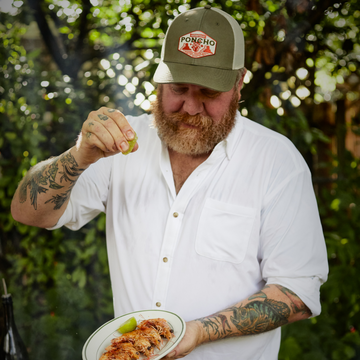 Bearded man in a white shirt and green cap squeezing lime over grilled shrimp on a plate outdoors