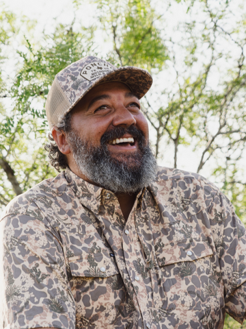 Smiling man wearing a patterned outdoor shirt and matching cap with trees in the background.