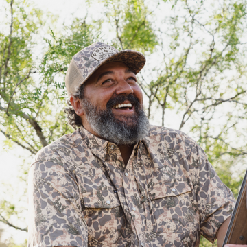 Smiling man wearing a patterned outdoor shirt and matching cap with trees in the background.