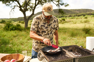 Man wearing a camouflage shirt cooks food in a cast iron skillet on an outdoor grill.
