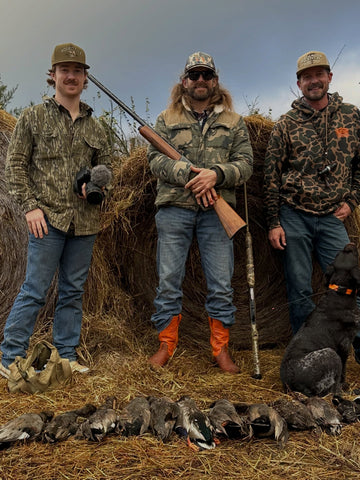 Three men in camouflage clothing pose outdoors with a rifle, hunting dogs, and harvested birds.