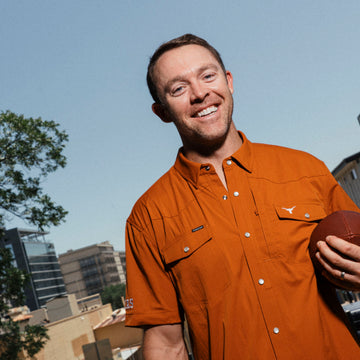 Smiling man in an orange button-up shirt holding a football outdoors against a city skyline