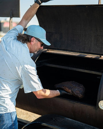Man in a blue cap and light shirt placing meat into an open outdoor smoker