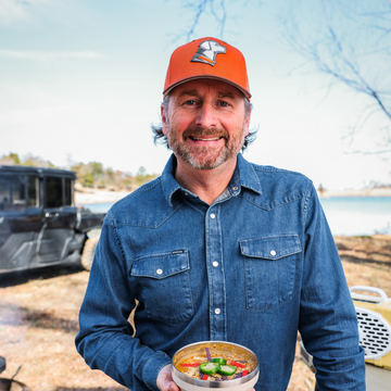 Man in a blue denim shirt and orange cap holding a bowl of soup outdoors near a lake with a vehicle in the background
