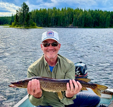 Man holding a large spotted fish on a boat with a forested lake in the background