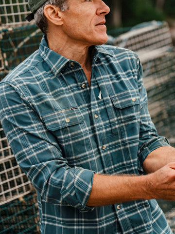 Man wearing a teal plaid outdoor shirt and green cap beside fishing nets