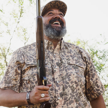Smiling bearded man in a camouflage shirt and cap holding a shotgun outdoors