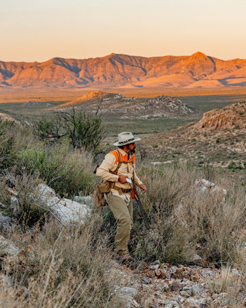 Person in a hat and orange vest walking through rocky desert brush with a mountain range in the distance