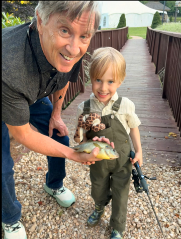 An adult and child smile while holding a fish beside a wooden dock, with a fishing rod nearby