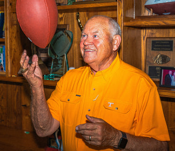 An older man wearing an orange shirt with a "T" logo is smiling and spinning a football on his finger indoors.