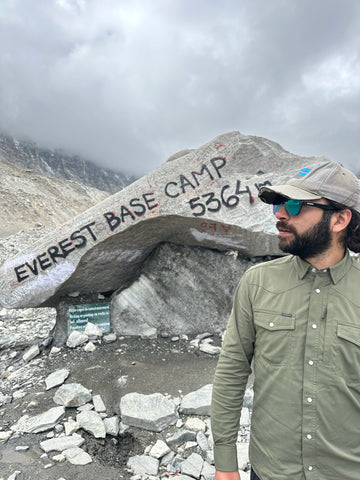 Man in an olive green outdoor shirt, sunglasses, and cap beside an Everest Base Camp marker rock