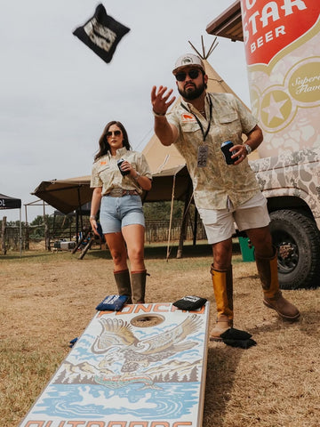 Man and woman in patterned outdoor button shirts at an outdoor event beside a cornhole board and beverage trailer