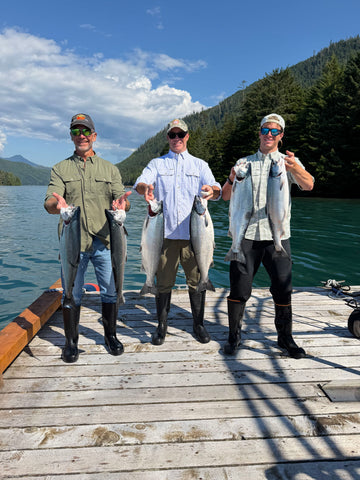 Three anglers in fishing attire hold fish on a wooden dock beside a mountain lake