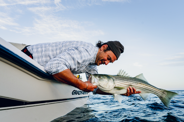 Man in a light plaid outdoor shirt holding a striped fish over ocean water from a boat