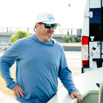Man wearing a blue long-sleeve shirt called The Spartina leaning on a boat outdoors by a van