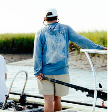 Man wearing The Spartina blue hoodie shirt standing on a boat steering with a natural marshland background