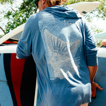 Man wearing a blue lightweight shirt with graphic design on back carrying a surfboard outdoors The Spartina