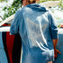 Man wearing a blue lightweight shirt with graphic design on back carrying a surfboard outdoors The Spartina