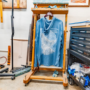 Blue long-sleeve shirt with a whale tail graphic and "Poncho Outdoors" text, hanging on an easel in a workshop setting called The Spartina.
