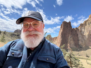 Bearded man in a blue outdoor shirt and cap posing before rugged rock formations under a blue sky