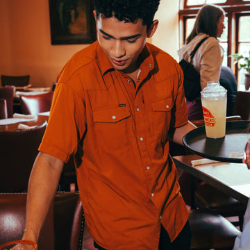 Young man wearing The 1969 orange short-sleeve shirt serving drinks in a lively cafe setting