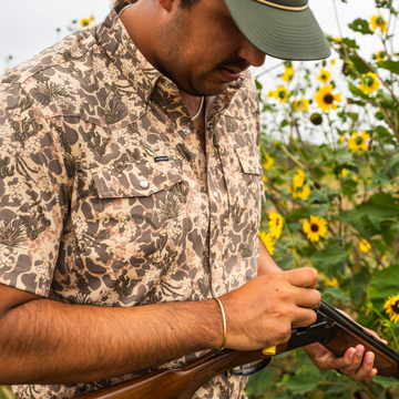 Man wearing The Abilene patterned shirt outdoors among sunflowers adjusting a wooden shotgun